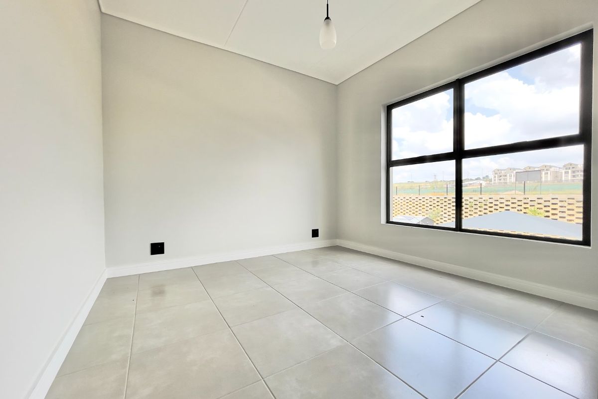 A bright, empty bedroom featuring neutral tiled flooring, modern black aluminium window frames, and a minimalist pendant light fixture.