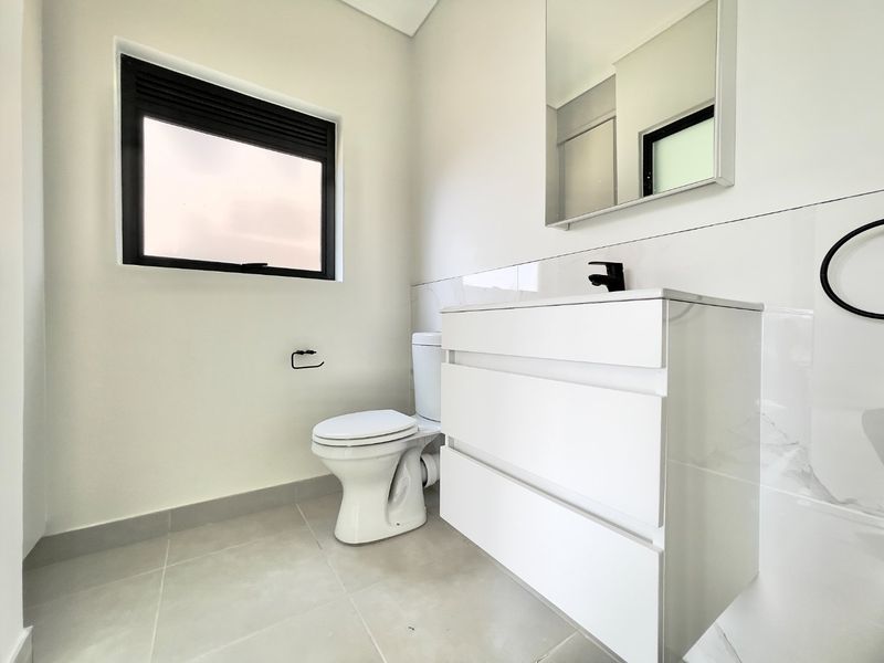 A modern, clean bathroom featuring a white vanity cabinet, basin, toilet, and wall-mounted mirror with neutral tiled flooring and wall finishes.