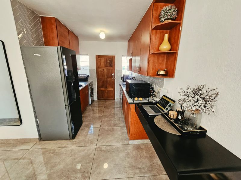 A functional galley-style kitchen featuring warm wood-toned cabinetry, herringbone backsplash tiling, and large format floor tiles, leading to an exterior door.