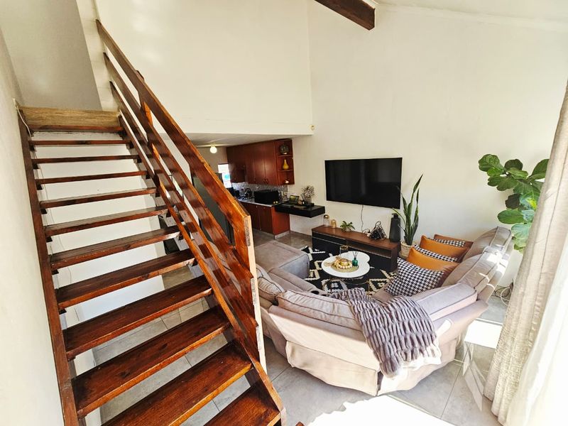 Open-plan living area featuring a wooden staircase, exposed ceiling beam, tiled floors, and a compact kitchen with built-in cabinetry.