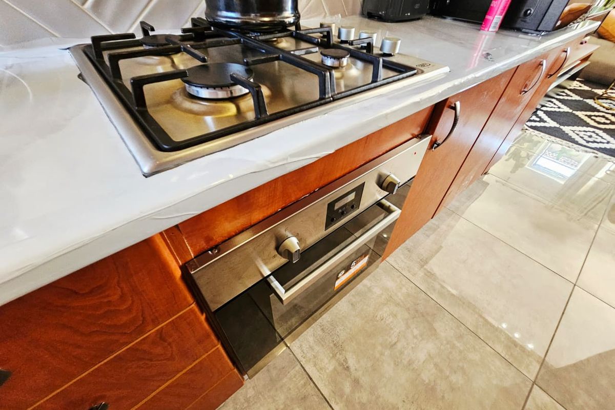 A modern kitchen featuring a gas hob, built-in oven, stone countertops, and stylish herringbone tile backsplash with warm wooden cabinetry.