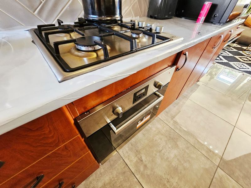 A modern kitchen featuring a gas hob, built-in oven, stone countertops, and stylish herringbone tile backsplash with warm wooden cabinetry.