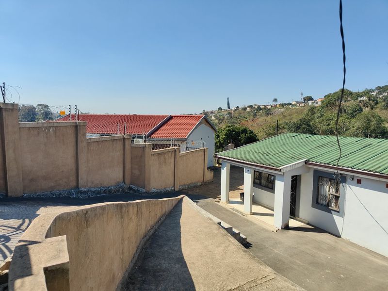 Exterior view of the property featuring a paved driveway, concrete boundary walls with security fencing, and a combination of tiled and corrugated metal roofing.