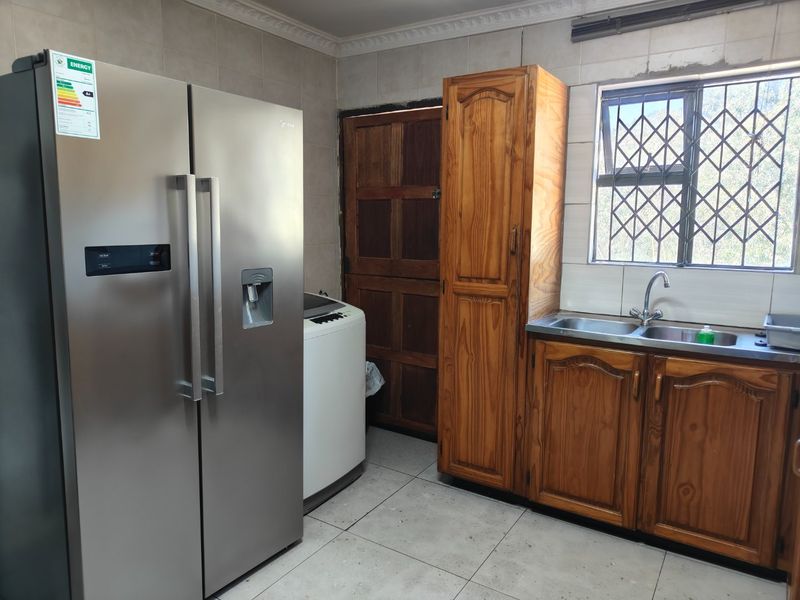 A functional scullery area featuring wooden cabinetry, a double stainless steel sink, tiled walls, and tiled flooring with a window security gate.
