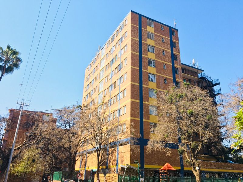 A multi-story apartment building featuring a brick facade with blue accents, surrounded by mature trees and security fencing.