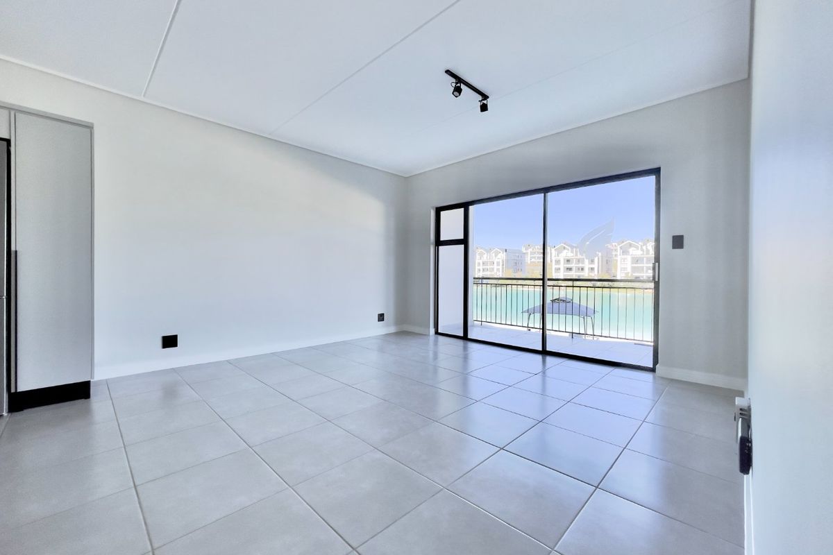 A bright, empty living room featuring neutral tiled flooring, modern track lighting, and a large sliding glass door leading to a balcony with water views.