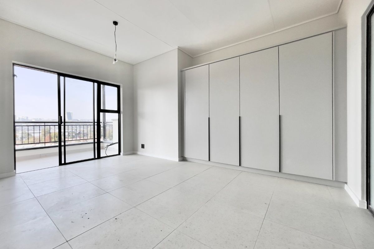 A bright, empty bedroom featuring modern built-in cupboards, large format floor tiles, and a sliding glass door leading to a private balcony.