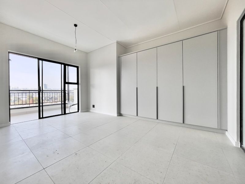 A bright, empty bedroom featuring modern built-in cupboards, large format floor tiles, and a sliding glass door leading to a private balcony.
