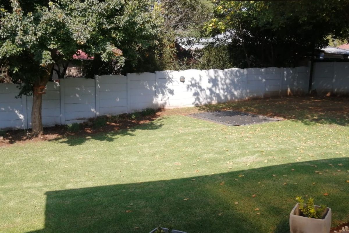 A view of the rear garden featuring a lawn, mature trees, and a precast concrete boundary wall, viewed from a covered tiled patio.