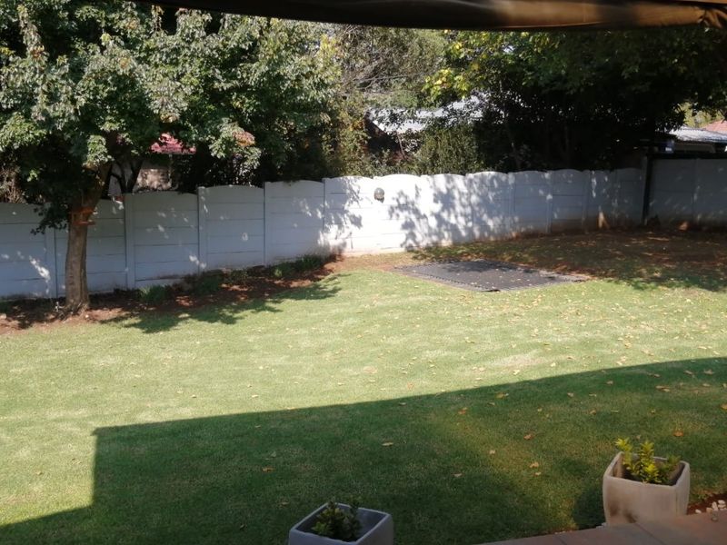 A view of the rear garden featuring a lawn, mature trees, and a precast concrete boundary wall, viewed from a covered tiled patio.