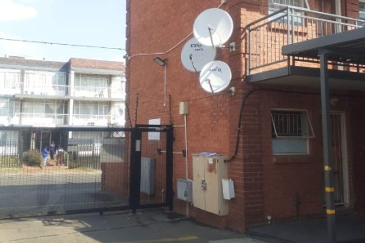 Exterior view of a brick apartment building featuring multiple satellite dishes, security fencing, and a gated entrance area.