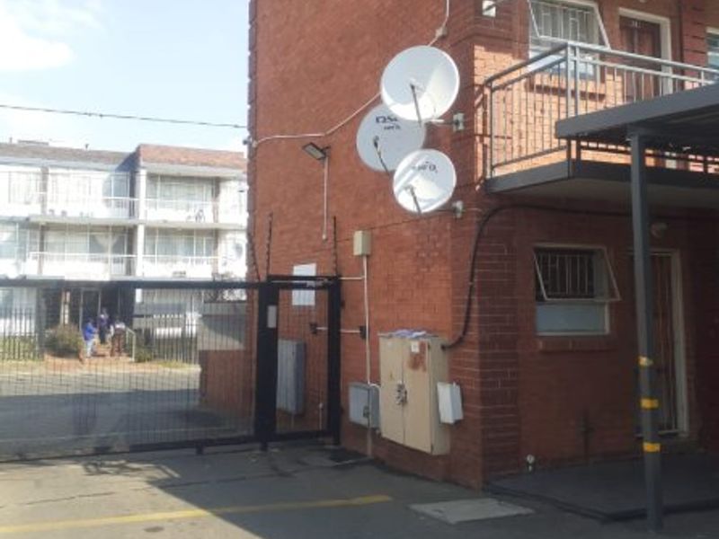 Exterior view of a brick apartment building featuring multiple satellite dishes, security fencing, and a gated entrance area.