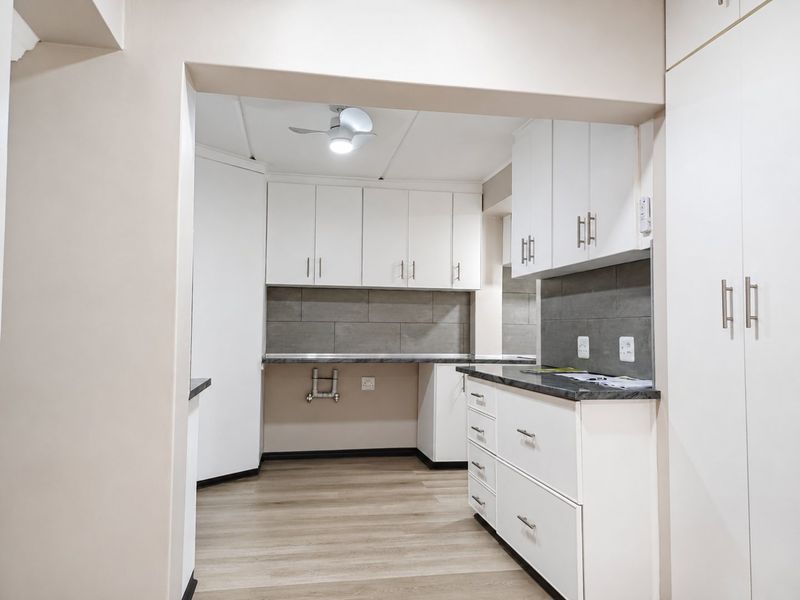 A clean, modern kitchen area featuring white built-in cabinetry, grey tiled backsplash, laminate countertops, and wood-look flooring.