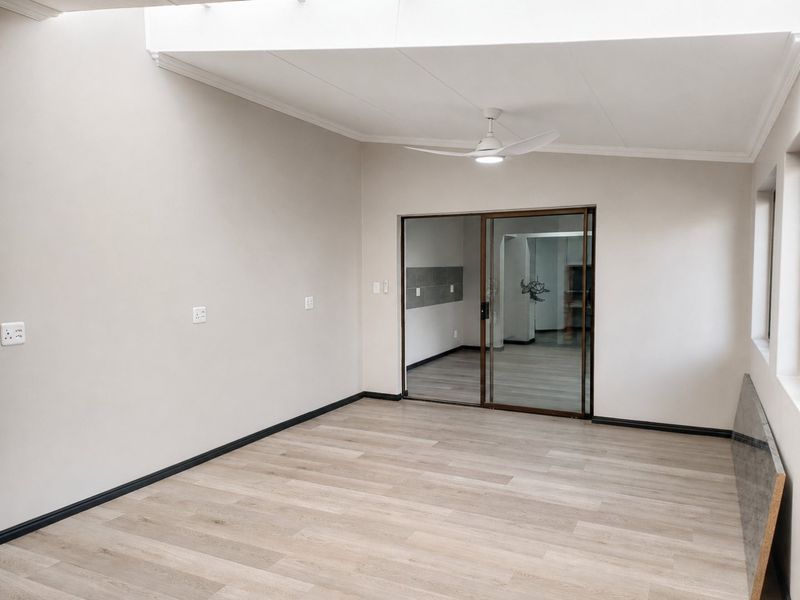 A bright, empty living area featuring modern laminate flooring, a ceiling fan, and a sliding glass door leading to an adjacent room.