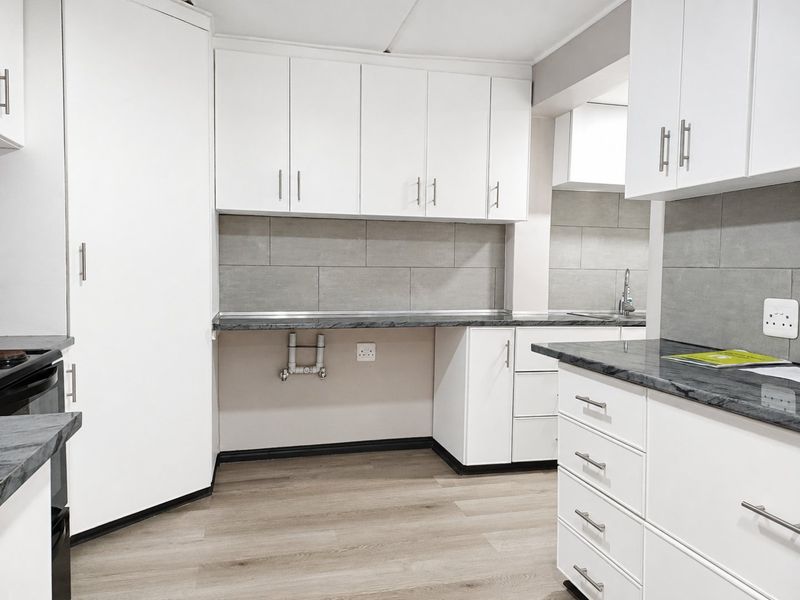 A clean, modern kitchen featuring white cabinetry, grey tiled backsplash, laminate countertops, and wood-look flooring, presented in a bright, empty space.