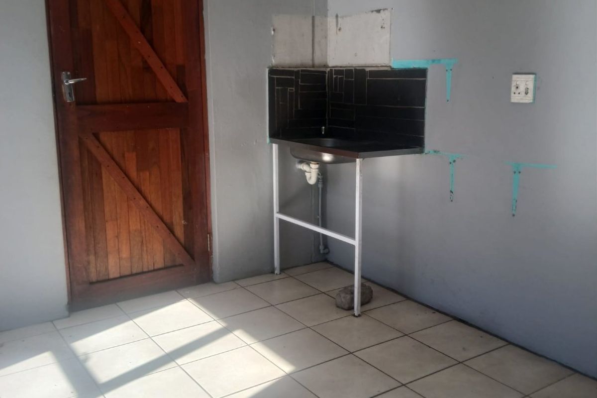 A basic, unfinished kitchen area featuring a simple stainless steel sink, tiled flooring, and a wooden door, requiring cosmetic updates and completion.