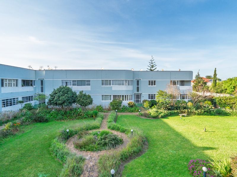 A view of the rear apartment building facade featuring a well-maintained communal garden, lawn, and brick pathways.