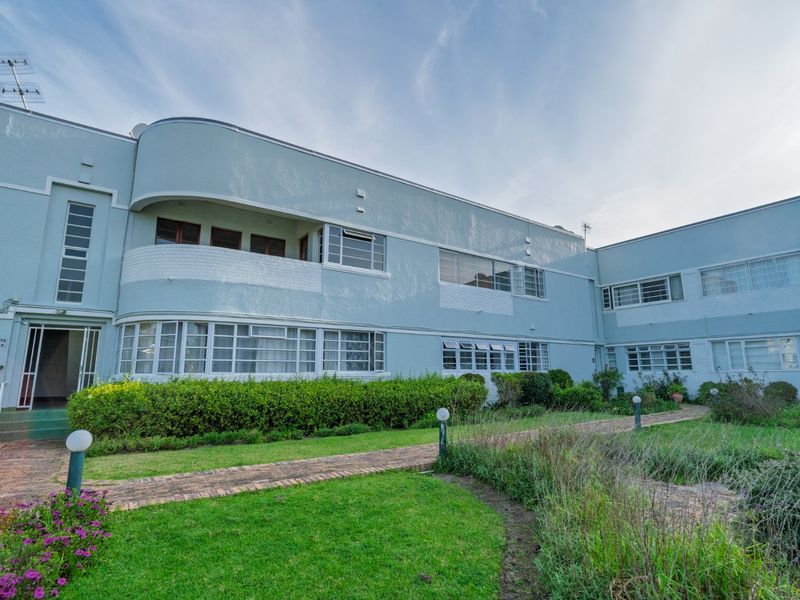Front view of an Art Deco style apartment building featuring a landscaped garden, brick pathways, and distinctive curved architectural balcony details.