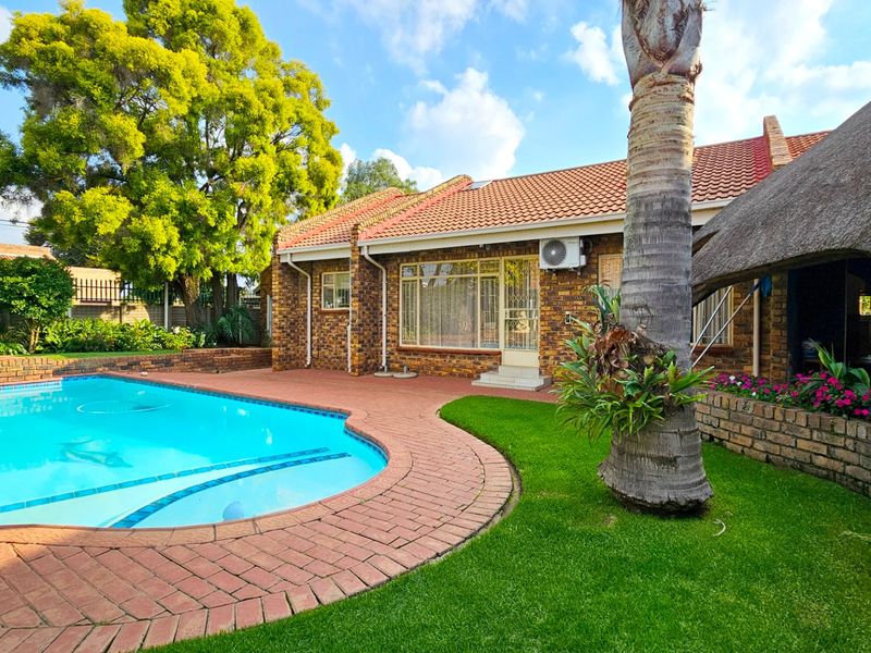 A well-maintained outdoor pool area featuring a swimming pool, brick paving, and a thatched roof structure set within a landscaped garden.