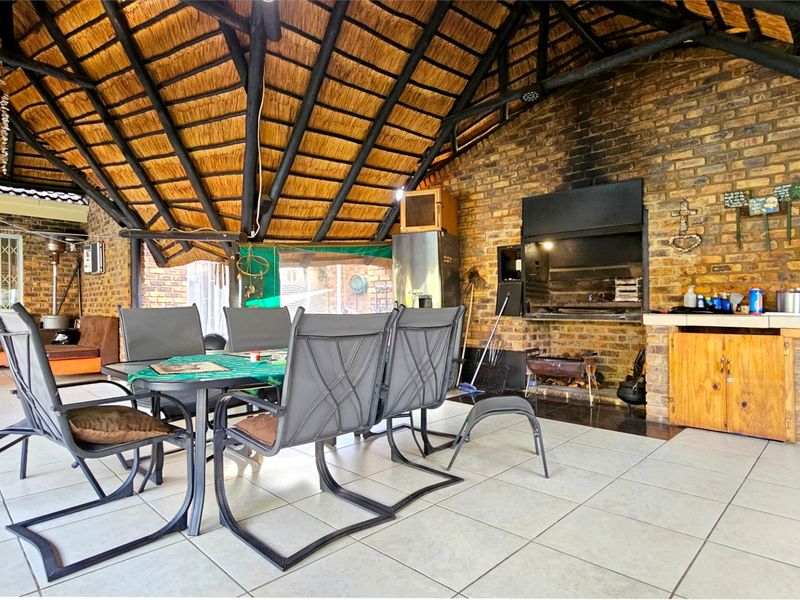 Spacious covered patio featuring a traditional thatched roof, exposed timber rafters, and a built-in brick braai area with tiled flooring.