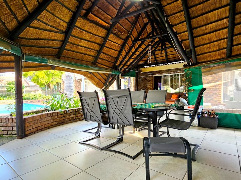 Covered outdoor patio featuring a traditional thatched roof, tiled flooring, and a brick retaining wall overlooking the garden and swimming pool area.