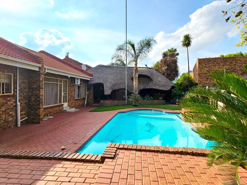Outdoor entertainment area featuring a swimming pool, brick paved patio, and a traditional thatched lapa set against a face brick house exterior.