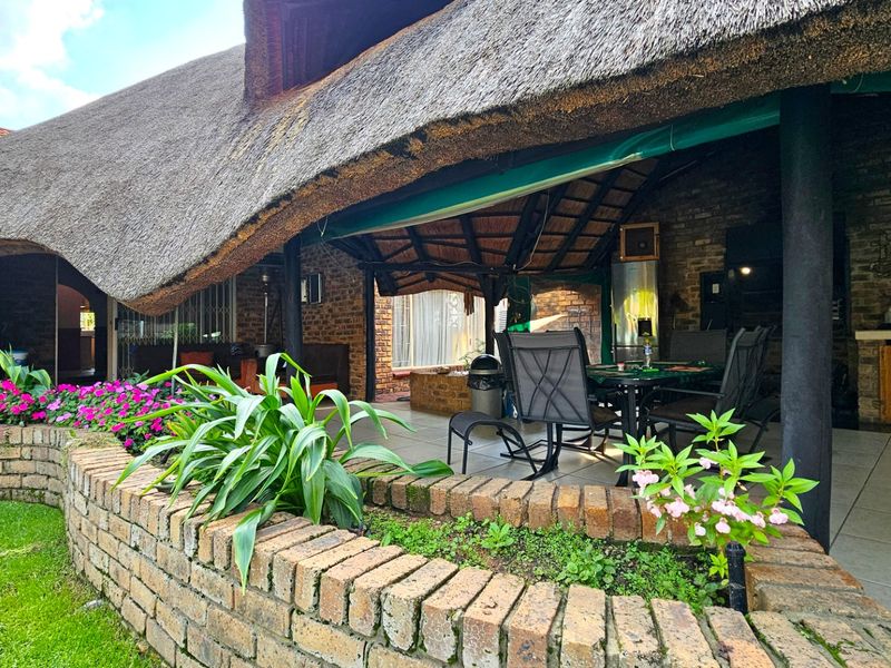 A spacious covered patio featuring a traditional thatched roof, brick masonry, and tiled flooring, overlooking a landscaped garden with a built-in brick planter.