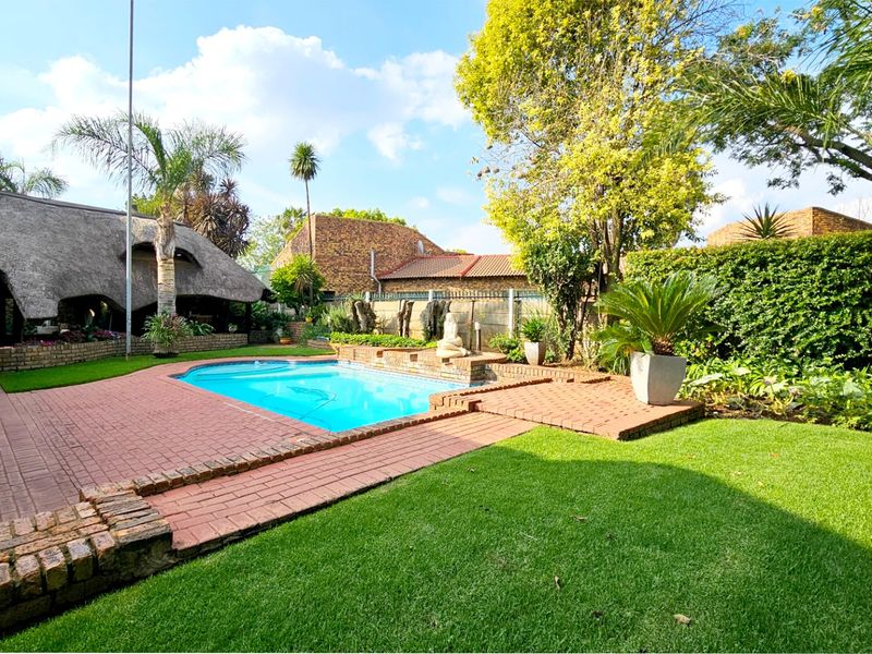 A well-maintained outdoor pool area featuring brick paving, a lush garden, and a covered thatched lapa, perfect for outdoor relaxation and entertainment.