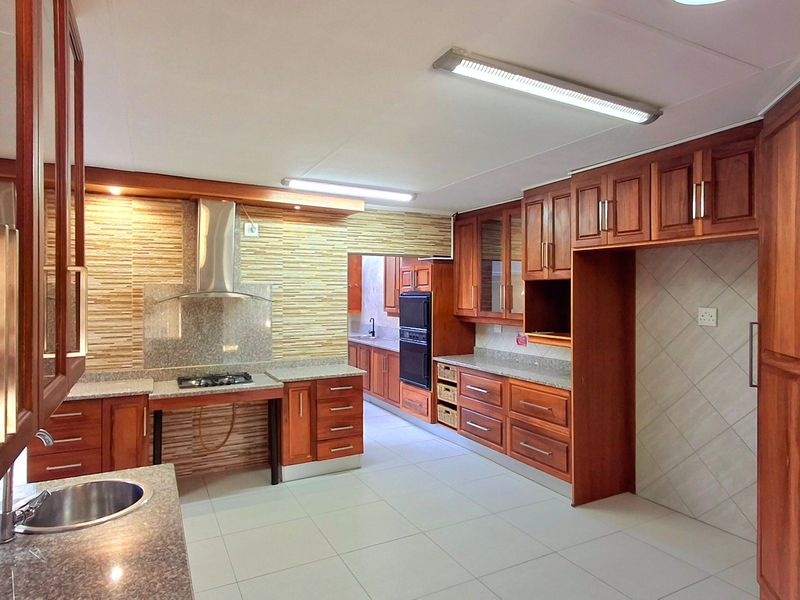 Spacious kitchen featuring extensive wooden cabinetry, granite countertops, a gas hob with extractor, and a built-in double oven, leading into a secondary prep area.