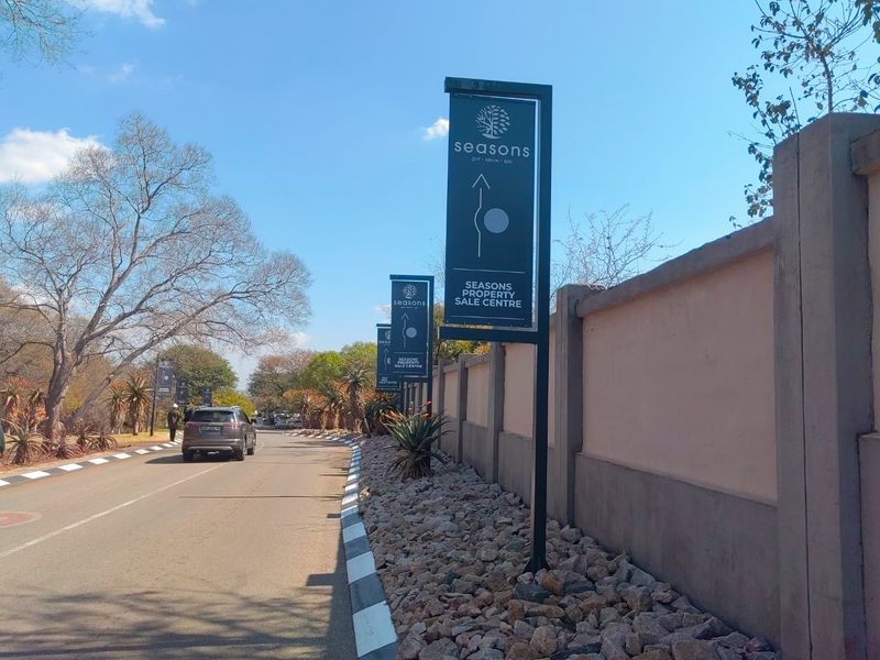 A view of the access road leading to the Seasons estate, featuring boundary walls and directional signage for the property sale centre.