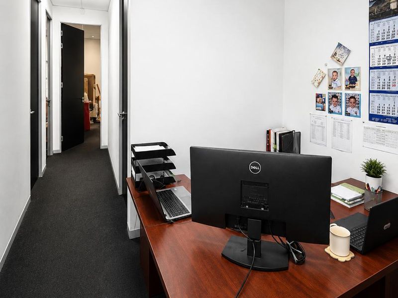 A functional office space featuring dark carpeting, a suspended ceiling with recessed lighting, and access to a connecting hallway.