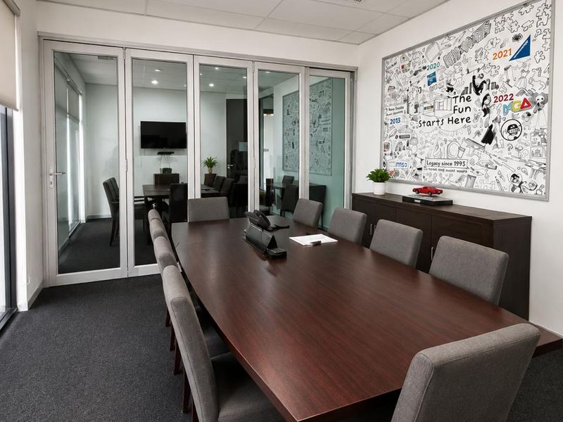 A professional boardroom featuring a dark wood conference table, grey upholstered chairs, and a glass partition wall overlooking an adjacent meeting space.