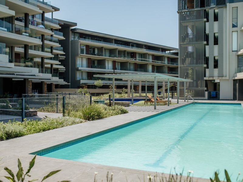 A well-maintained communal swimming pool area featuring a modern pergola, paved surround, and landscaped gardens within a contemporary apartment complex.