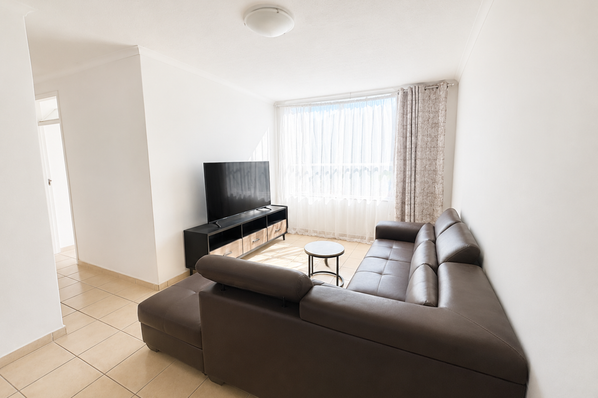 A bright living room featuring neutral tiled flooring, white walls, and a large window with sheer curtains, providing a clean and functional living space.