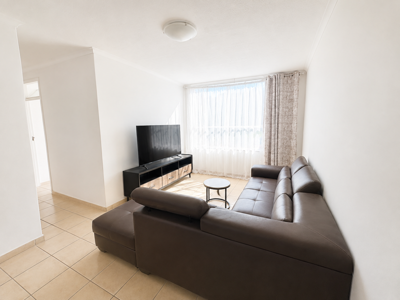 A bright living room featuring neutral tiled flooring, white walls, and a large window with sheer curtains, providing a clean and functional living space.