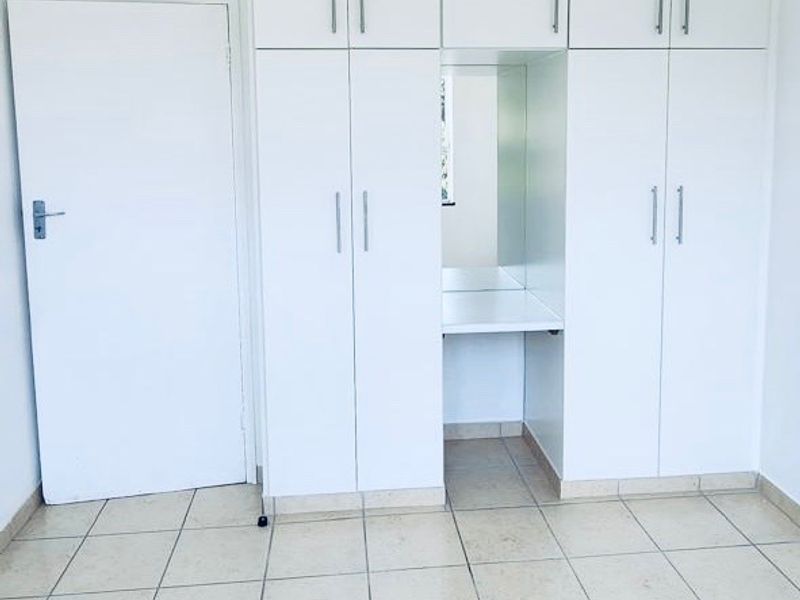 A bright, empty bedroom featuring white built-in cupboards with an integrated vanity area and neutral tiled flooring.