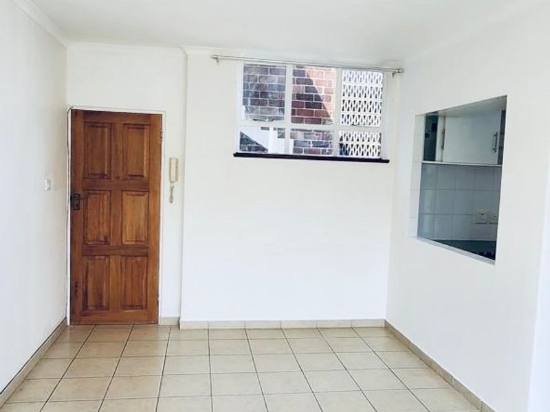 An empty living area featuring tiled flooring, a solid timber door, and a convenient pass-through window connecting to the kitchen space.