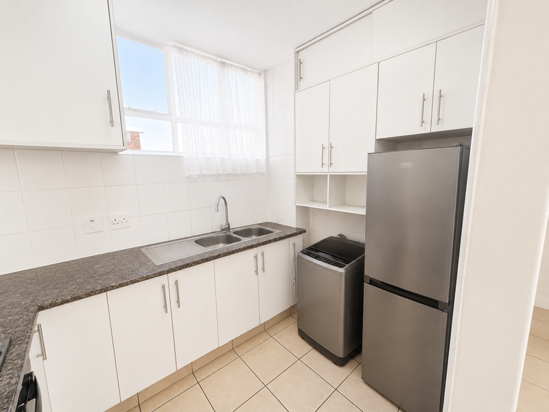 A functional kitchen featuring white built-in cabinetry, dark laminate countertops, a stainless steel sink, and tiled flooring with ample storage space.