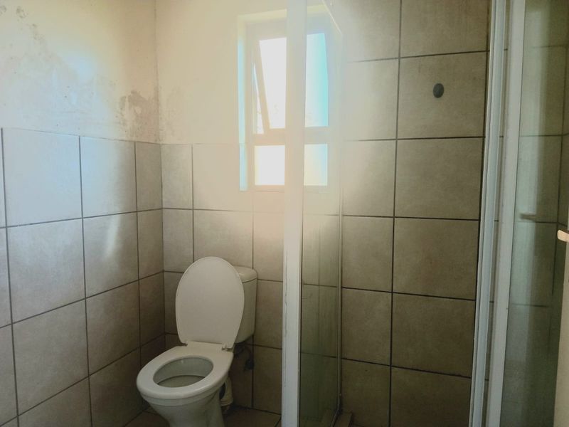 A compact bathroom featuring a toilet and shower enclosure with tiled walls, currently showing signs of wall surface wear and peeling paint.
