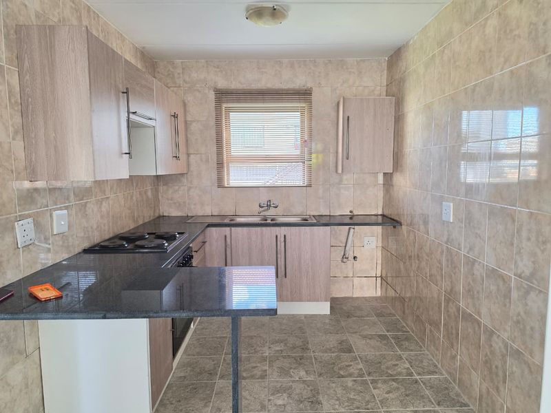 A functional kitchen featuring light wood-finish cabinetry, dark granite countertops, an electric hob, built-in oven, and fully tiled wall surfaces.