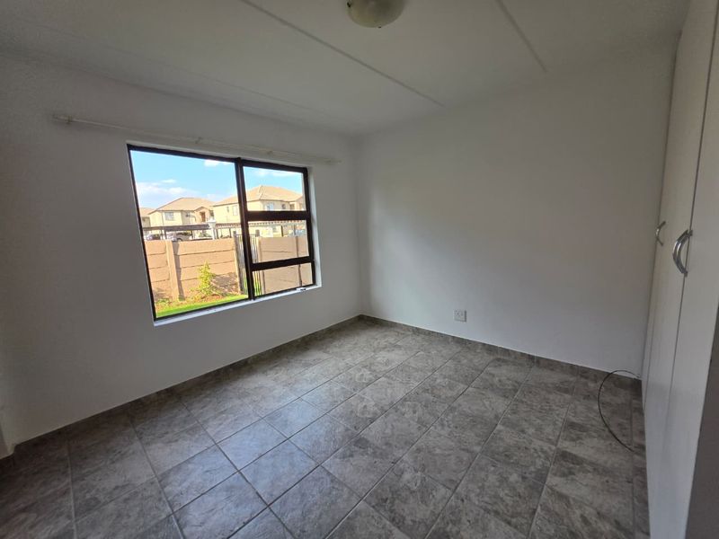 A bright, empty bedroom featuring tiled flooring, built-in cupboards, and a large window providing natural light and a view of the surrounding complex.