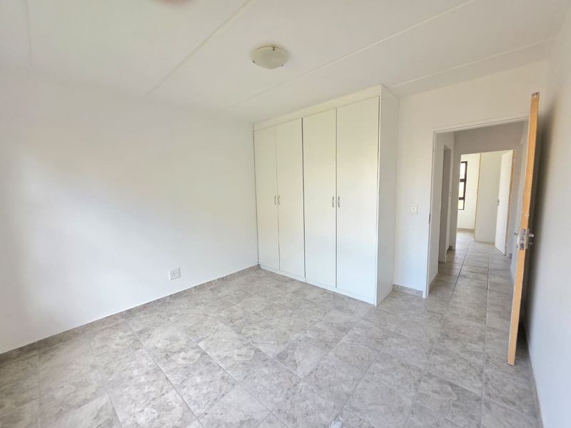 A bright, empty bedroom featuring neutral tiled flooring and ample built-in cupboard storage, leading into a hallway.
