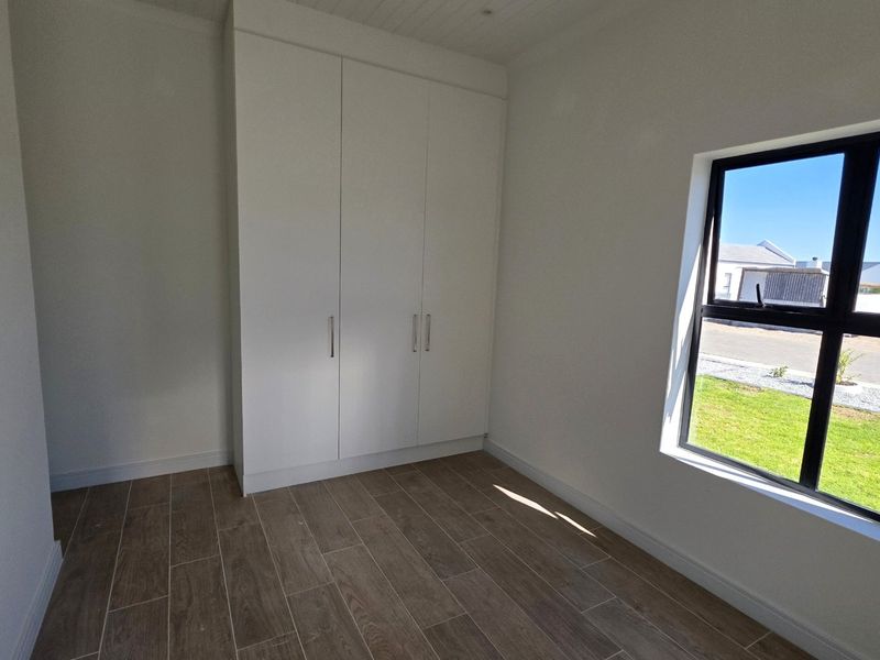 A bright, empty bedroom featuring modern wood-look tiled flooring, built-in cupboards, and a large window with an exterior view.