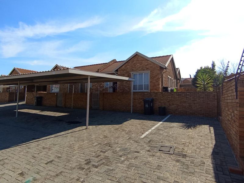 Front exterior view of a brick townhouse featuring a covered carport, paved driveway, and secure perimeter wall with electric fencing.