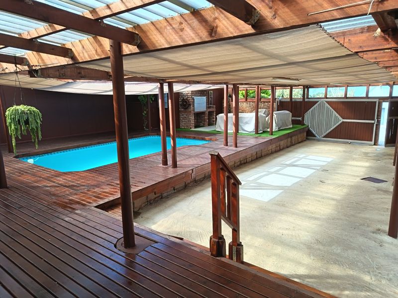 Covered outdoor entertainment area featuring a swimming pool, extensive timber decking, and a built-in braai under a timber pergola structure.