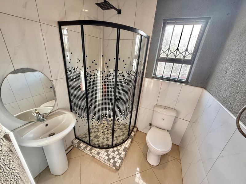 A modern bathroom featuring a corner shower enclosure, pedestal basin, and toilet with white tiled walls and neutral floor tiles.