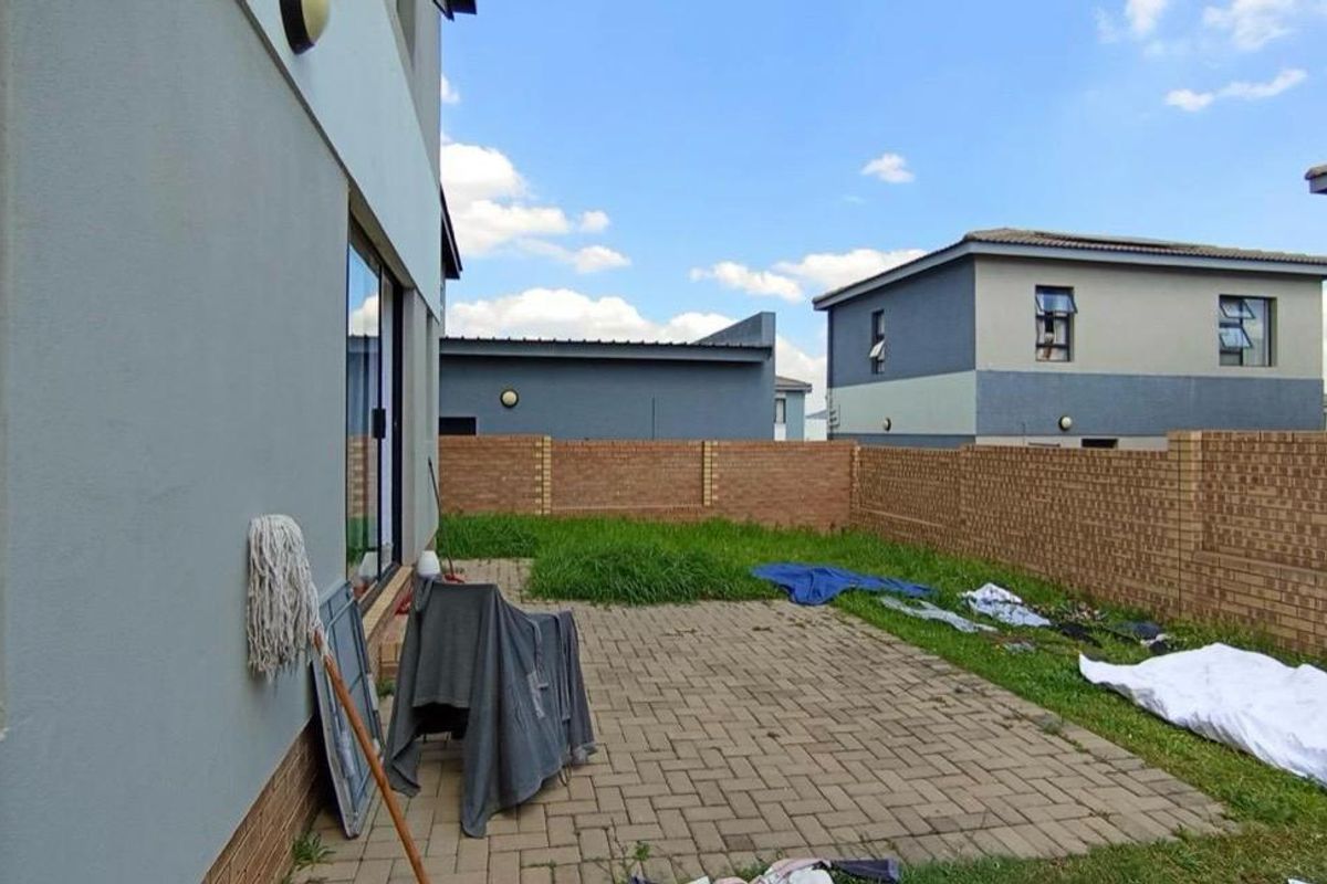 A rear exterior view of the property featuring a paved patio area, a small lawn, and a brick boundary wall.