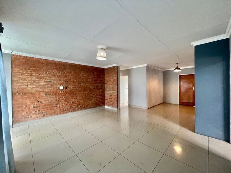 Spacious, empty living area featuring an exposed brick feature wall, neutral tiled flooring, and decorative ceiling cornices.