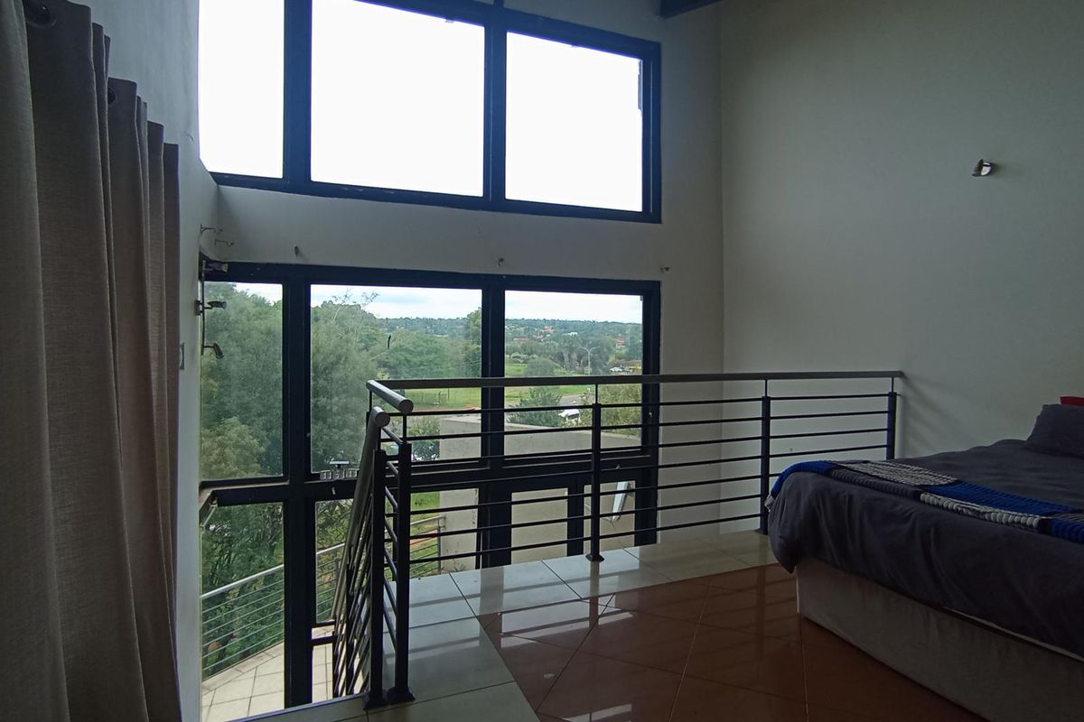 A mezzanine bedroom featuring double-volume windows, tiled flooring, and a modern metal balustrade overlooking the surrounding landscape.