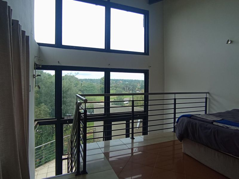 A mezzanine bedroom featuring double-volume windows, tiled flooring, and a modern metal balustrade overlooking the surrounding landscape.